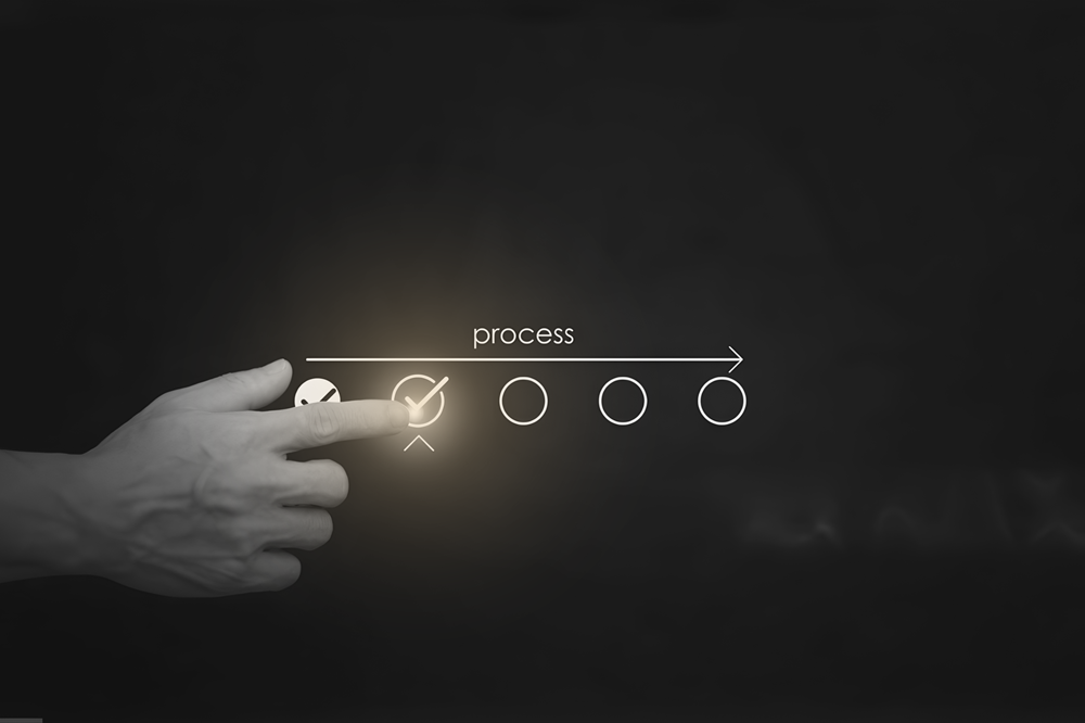 Hand points to the completed first step of a four-step process diagram on a dark chalkboard background. The first circle is checked, indicating completion.
